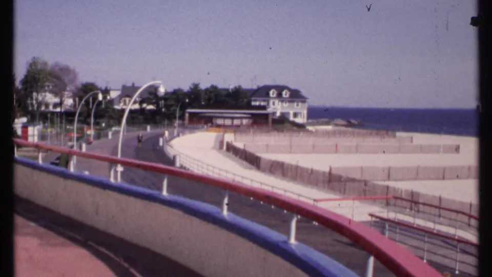 1969 home-movie frame from New London showing a boardwalk railing sweeping past beach fencing, a coastal road, houses, trees, and ocean.