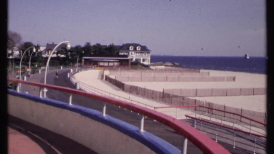 1969 New London coastal view with red and blue boardwalk railings, fenced beach, small road, shoreline buildings, trees, and ocean horizon.