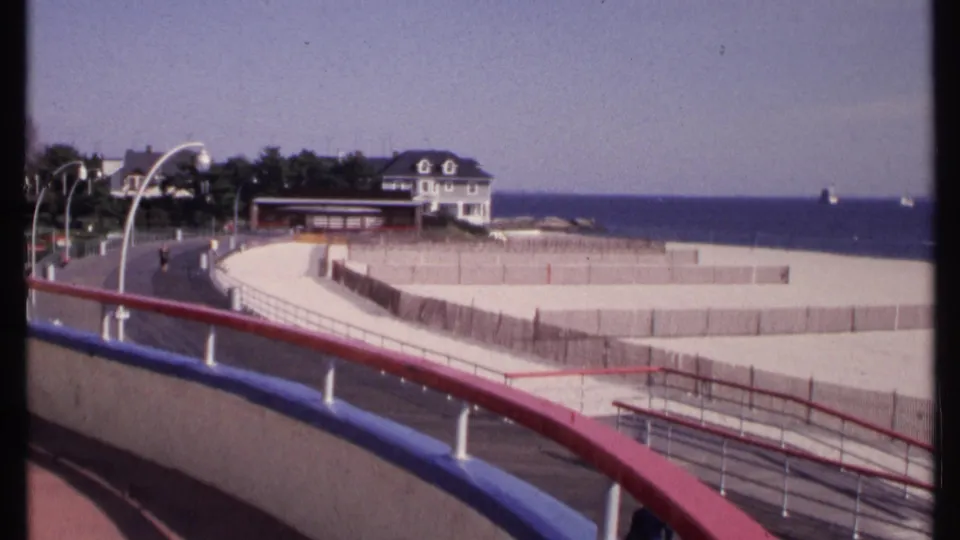 1969 New London coastal boardwalk view with painted railings, arched lamps, beach fencing, ocean, and a white house near trees.