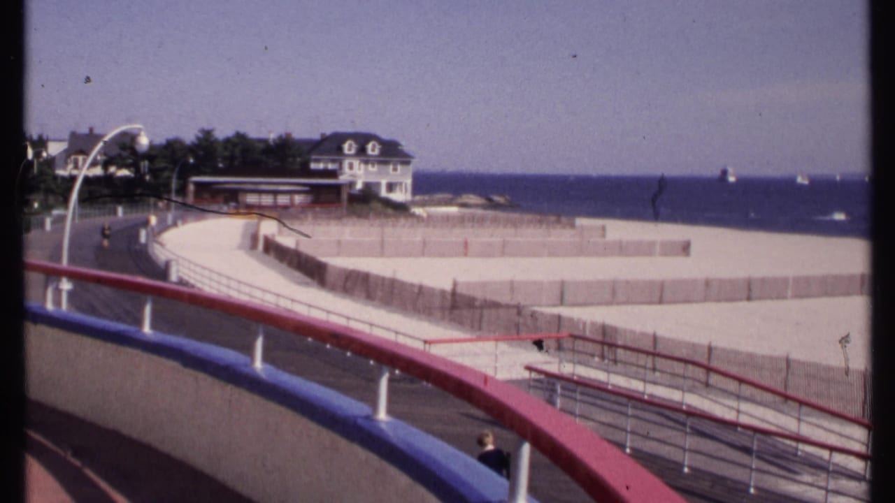 Seaside promenade with painted railings, beach fencing, ocean horizon, white house, and small pedestrians.