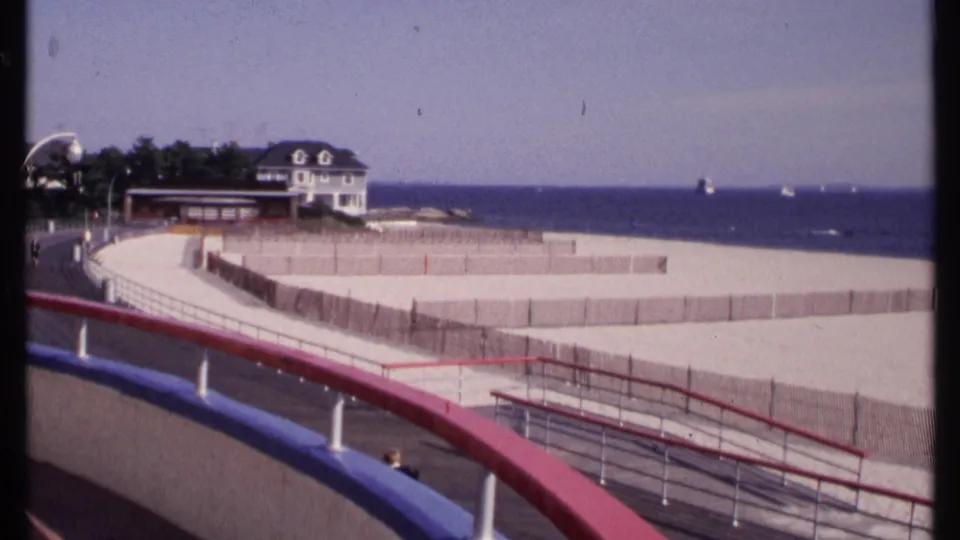 New London, Connecticut boardwalk view from 1969 with red and blue railings, fenced sand, trees, a white house, and sailboats offshore.