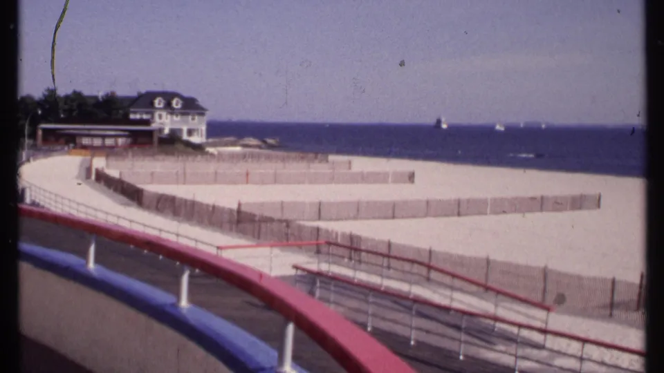 1969 New London waterfront scene with curved promenade railings, fenced sand, distant pedestrians, a white house, trees, and sailboats.
