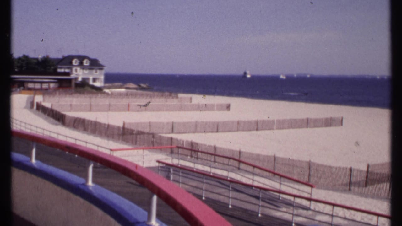 1969 New London beach view from a boardwalk with red and blue railings, wooden sand fencing, a beach house, ocean, and boats on the horizon.
