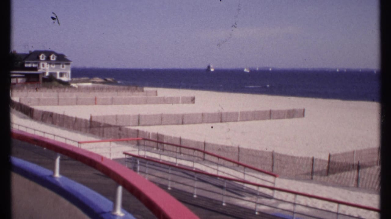 New London, Connecticut beach scene with boardwalk railings in the foreground, fenced sand, a beach house at left, and distant boats offshore.