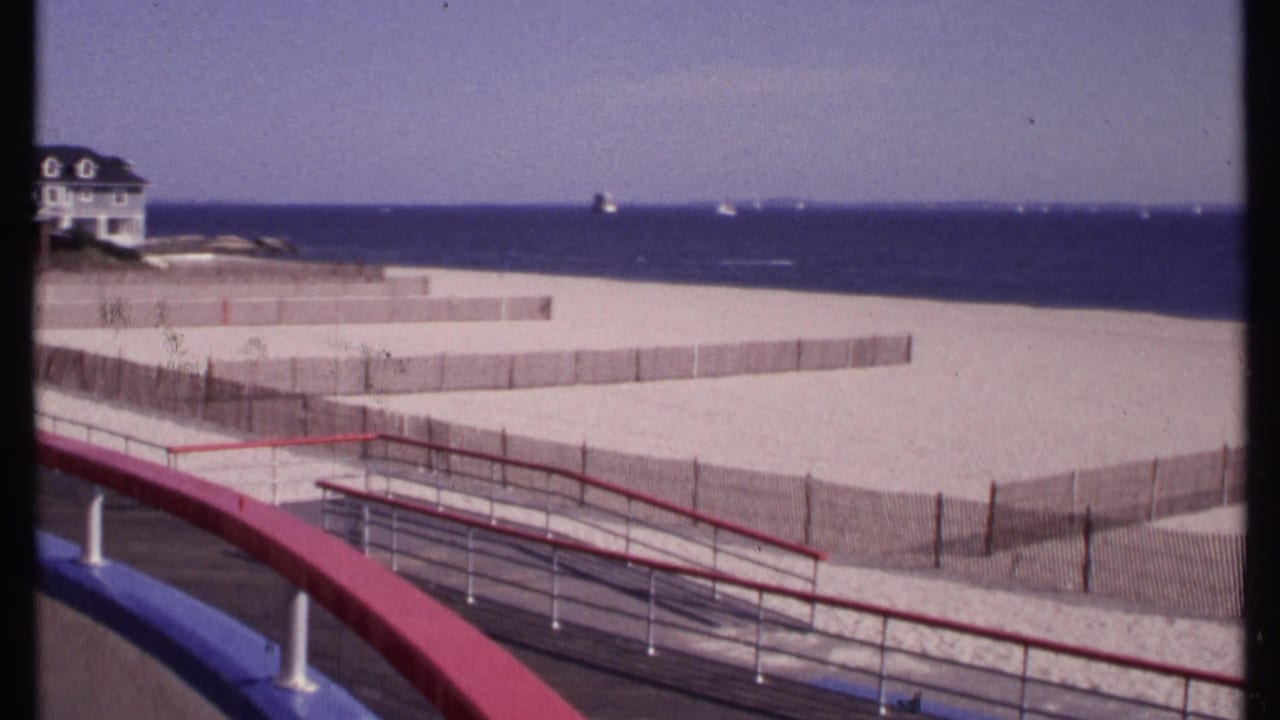 1969 home-movie frame looking across New London beach fencing from a boardwalk, with red and blue railings and the ocean horizon beyond.