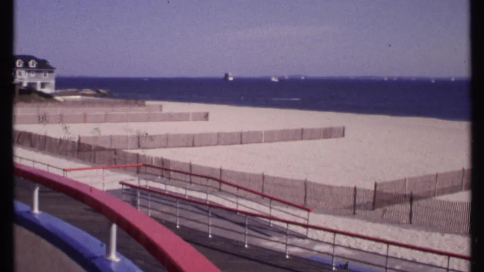 1969 home-movie frame looking across New London beach fencing from a boardwalk, with red and blue railings and the ocean horizon beyond.