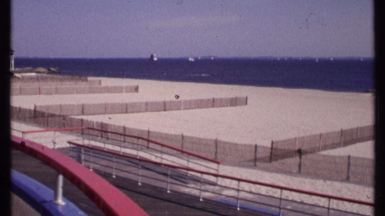 New London beach view on 8mm film, showing foreground boardwalk rails, wooden sand fencing, open beach, blue ocean, and small distant boats.