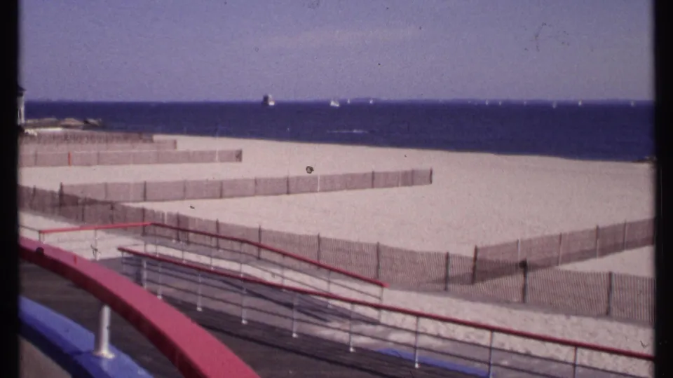 New London beach view on 8mm film, showing foreground boardwalk rails, wooden sand fencing, open beach, blue ocean, and small distant boats.