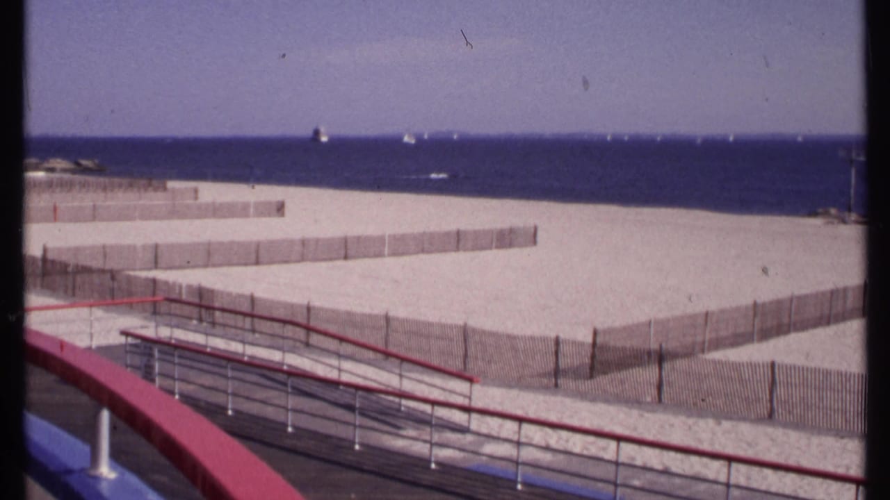 1969 New London beach view from a balcony, with wooden sand fencing, ocean horizon, distant boats, and foreground railings.