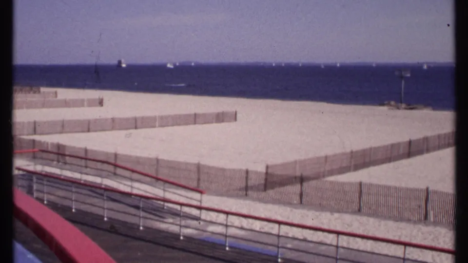Balcony view of a divided New London beach in 1969, with pale sand, low fencing, ocean water, and small boats offshore.
