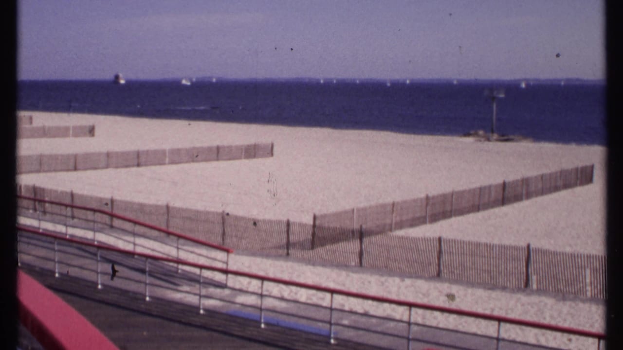 1969 elevated home-movie frame of New London beach fencing, blue water, a foreground walkway, and a tall shoreline post.