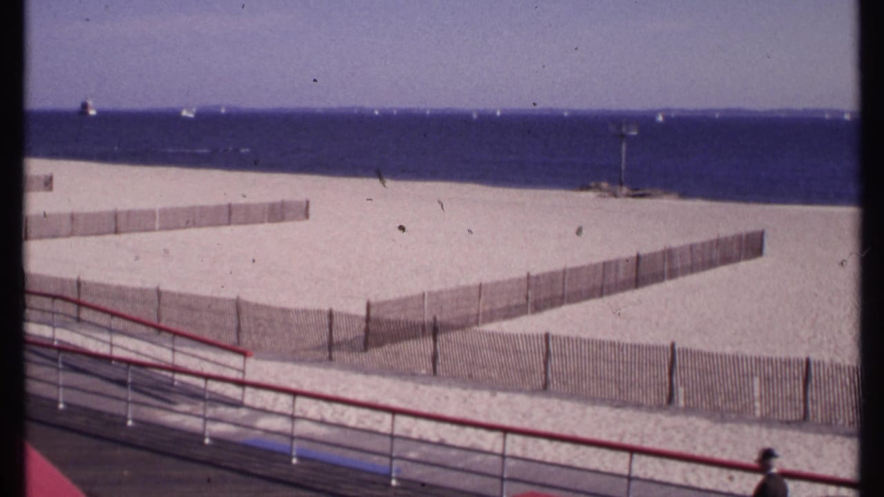 1969 New London beach view with sand fencing, distant sailboats, blue ocean, and a red boardwalk railing in the foreground.