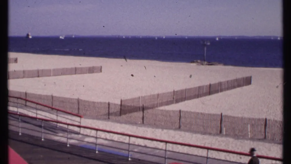 1969 New London beach view with sand fencing, distant sailboats, blue ocean, and a red boardwalk railing in the foreground.