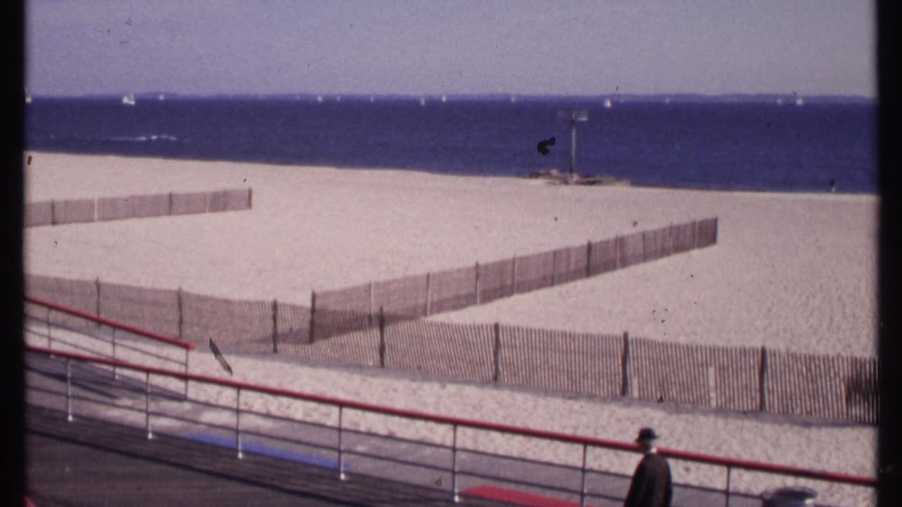 A man in dark clothing passes along the foreground boardwalk as the 1969 New London beach and ocean remain in view.