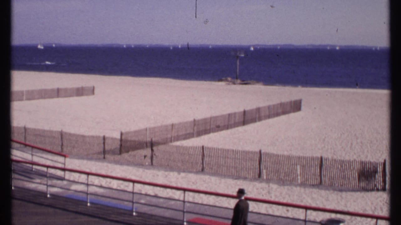 New London beach scene with red railing, wooden fencing, blue ocean, and an unidentified man at the lower edge of the frame.