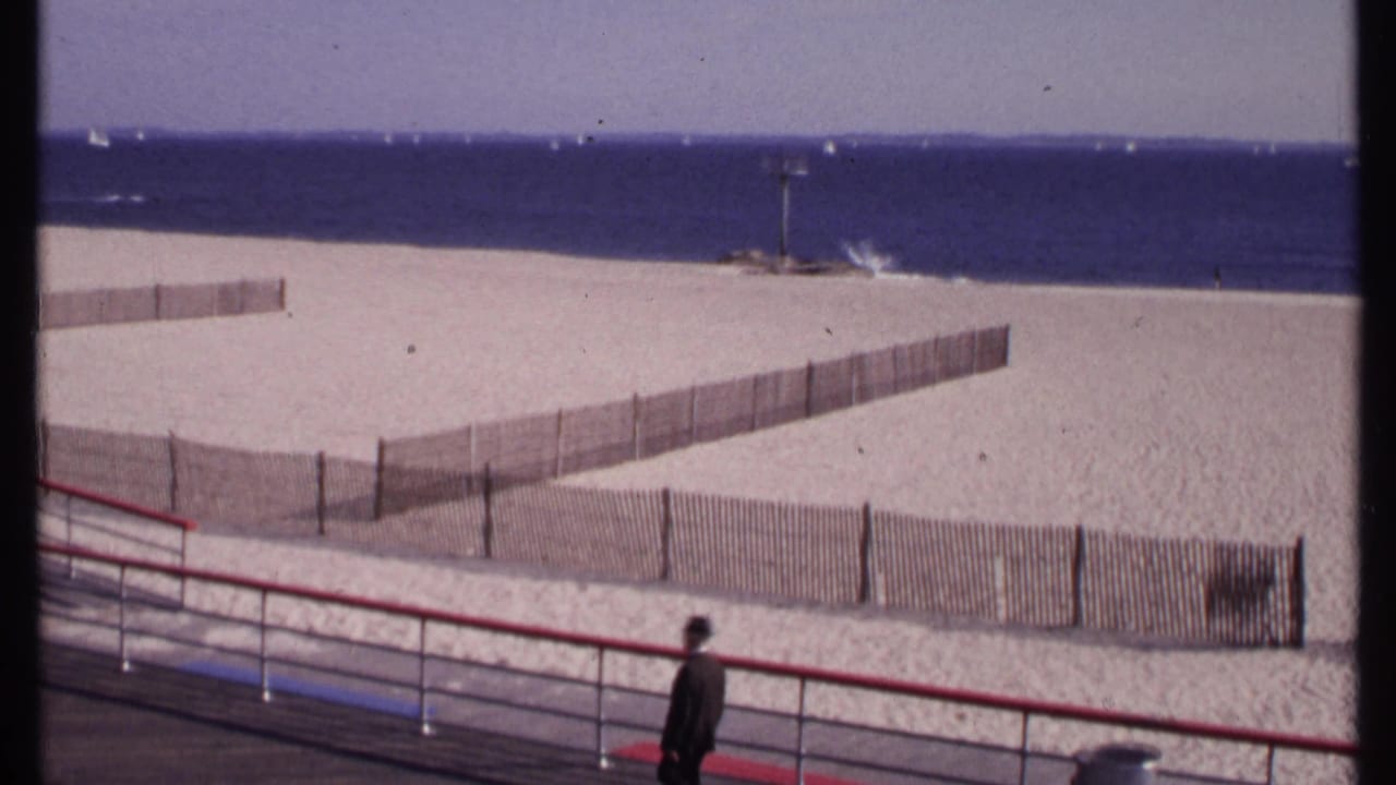 A man in dark clothing stands or walks near the red railing with the 1969 New London beach, fencing, and ocean behind him.