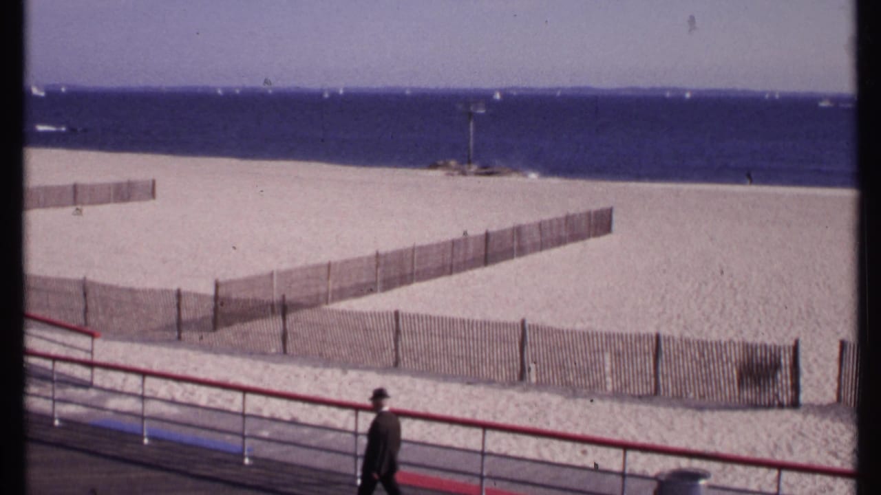 Man in a dark suit and hat walks along a boardwalk above a sandy beach and ocean.