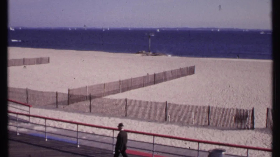 1969 home-movie frame from New London, Connecticut showing a suited man on a boardwalk above beach fencing, sand, and ocean.