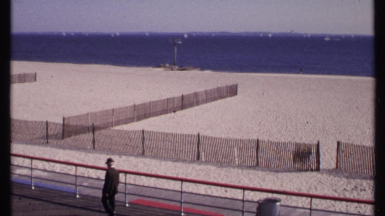 Man in dark clothing passes a red boardwalk rail with beach fencing and ocean behind him.
