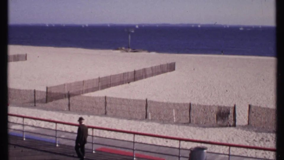 A distant suited man in a hat walks past the red railing on a New London beach boardwalk in 1969 home-movie footage.