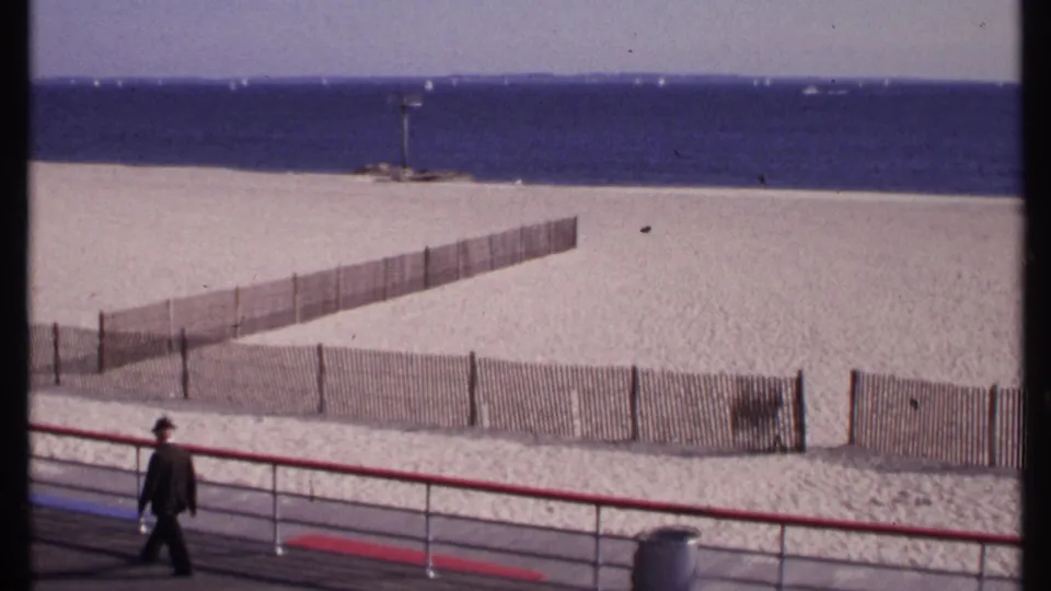 The man nears the edge of frame while the beach fence, broad sand, and blue water dominate the New London shoreline view.
