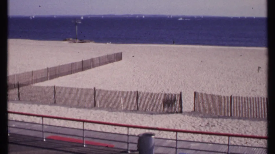 A New London beach view after the man has left the frame, with red railing, wood fencing, sand, ocean, and distant boats visible.
