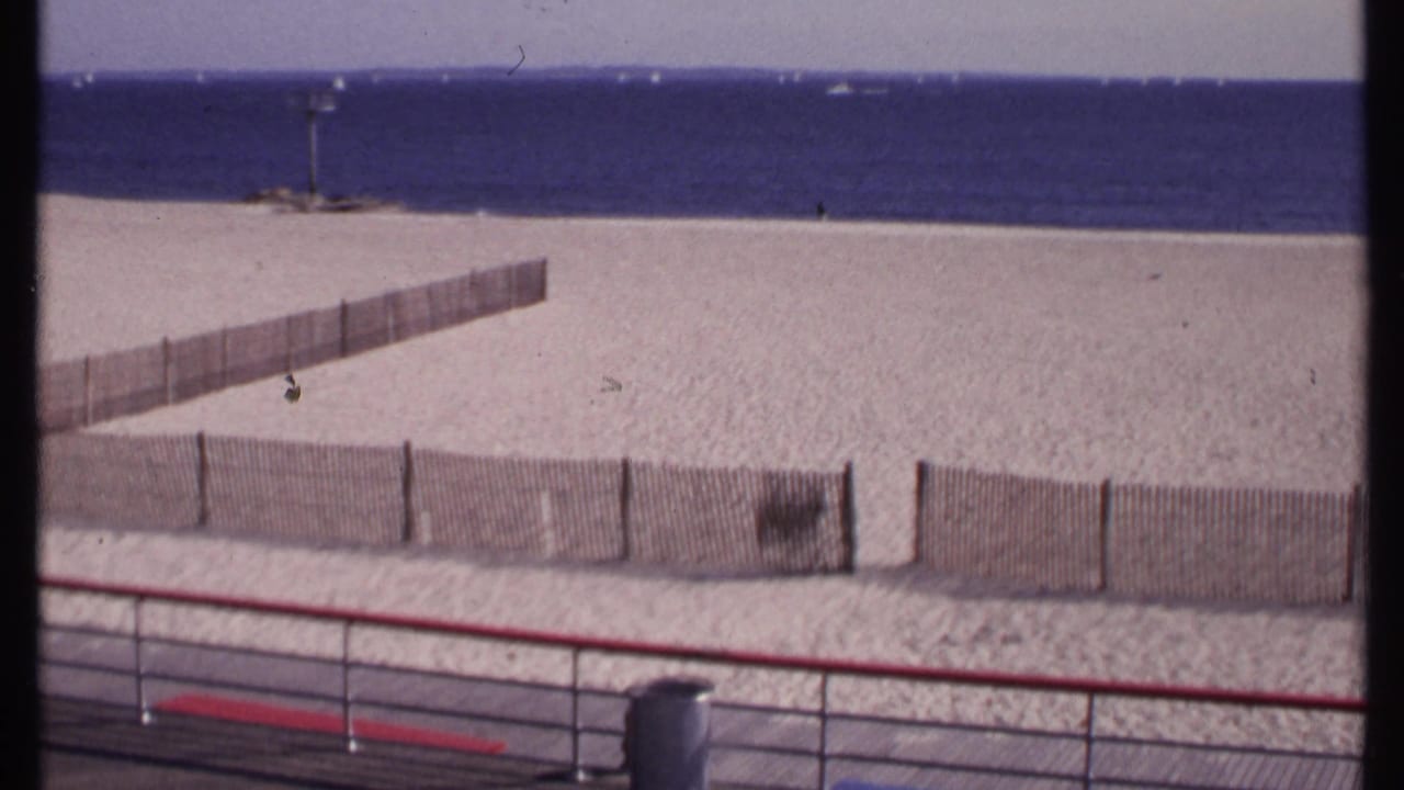 Boardwalk view over a fenced sandy beach toward blue water and distant small boats.