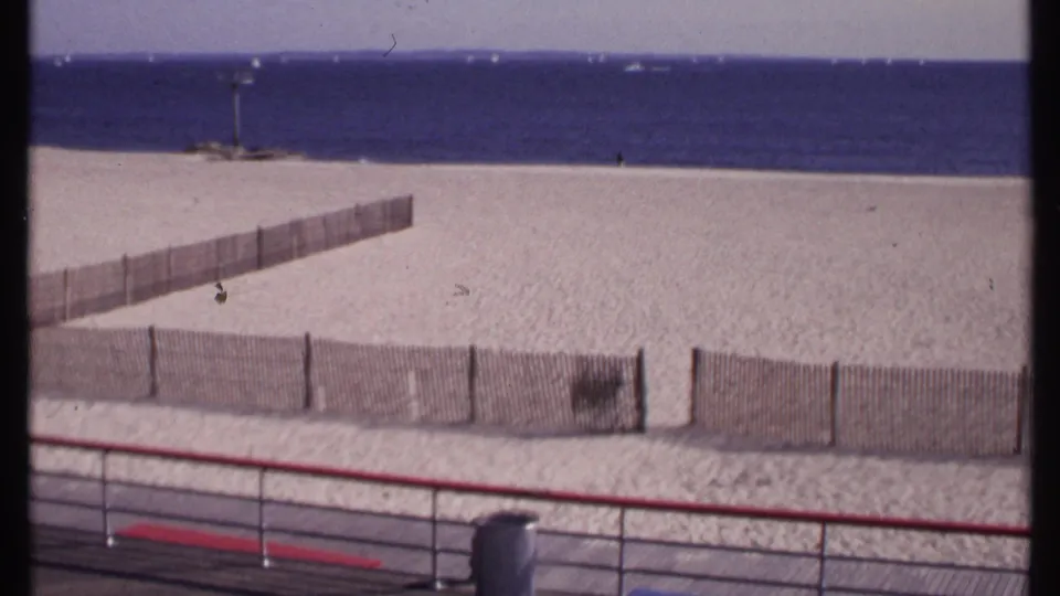 1969 New London home-movie frame showing a quiet fenced beach, boardwalk railing, trash can, blue water, and distant boats.