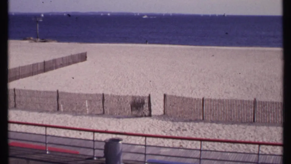1969 New London frame with wooden beach fencing, pale sand, a red boardwalk rail, calm water, and small boats near the horizon.