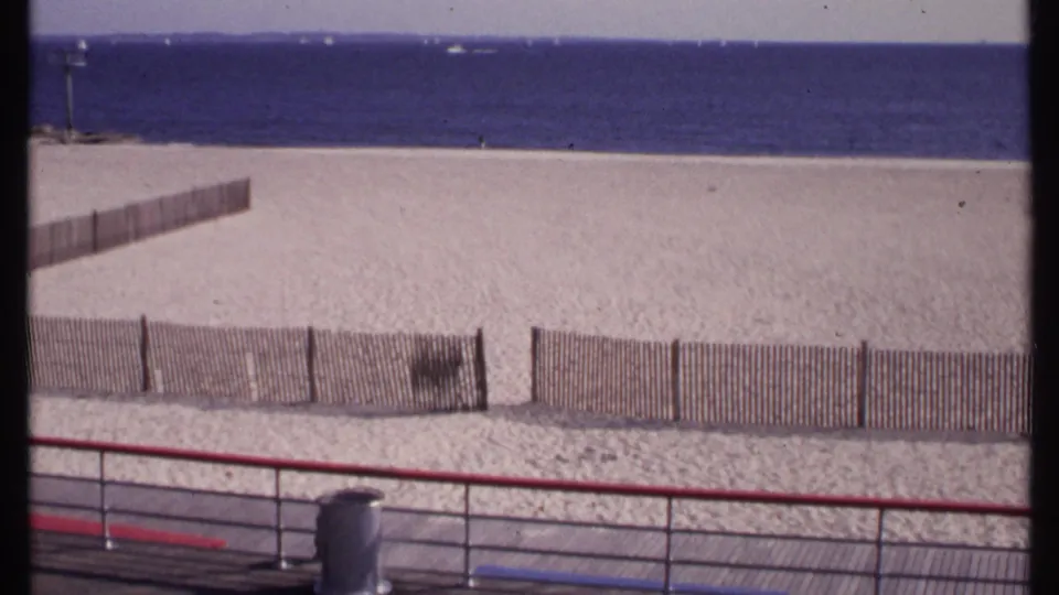 1969 New London beach view from a boardwalk, showing fencing across pale sand, blue ocean, and a steady home-movie composition.