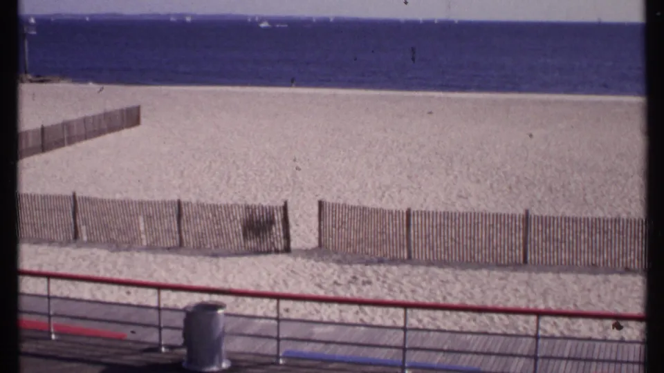 1969 New London coastal frame with a red-railed boardwalk foreground, pale fenced beach, blue water, and tiny boats offshore.
