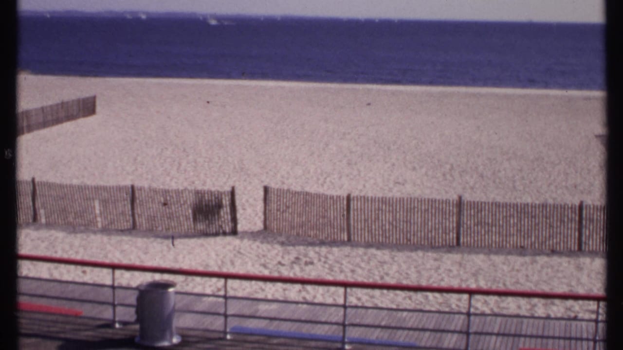 Boardwalk view over a fenced sandy beach with calm water beyond.