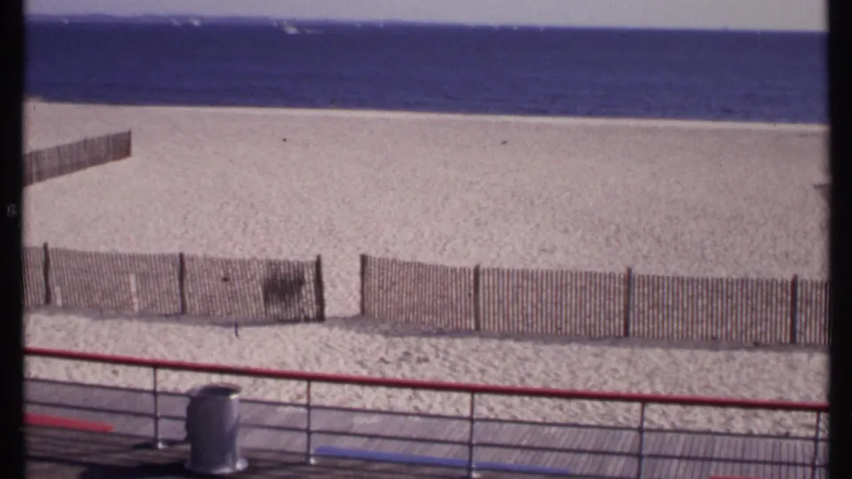 1969 New London beach view from a boardwalk, with low wooden fencing, pale sand, calm water, and a distant shoreline horizon.