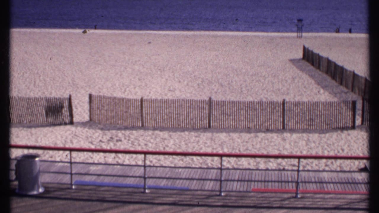 New London beach in 1969 with low slat fencing, boardwalk railings, calm blue water, and a small lifeguard stand in the distance.
