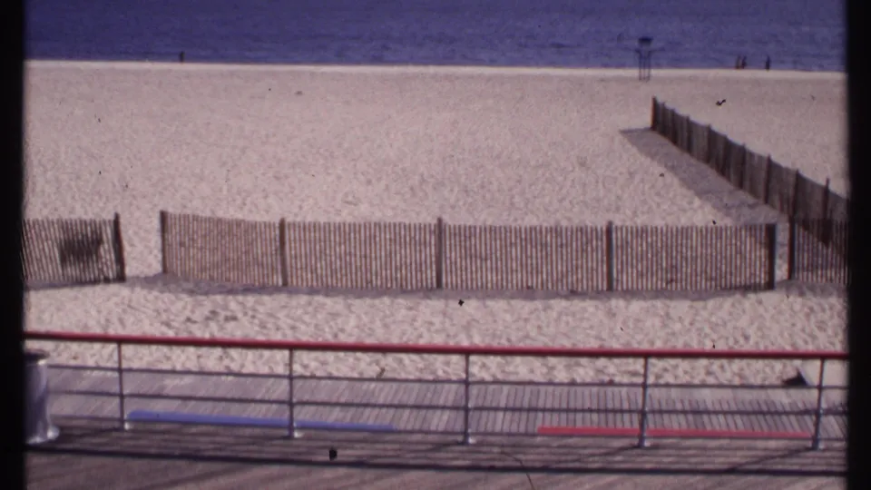 1969 coastal beach scene in New London, showing a boardwalk overlook, low fence panels, open sand, calm water, and tiny distant visitors.