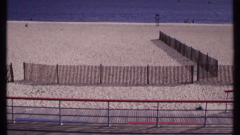 New London, Connecticut beach view from 1969 with boardwalk railing, low wooden fencing, pale sand, calm water, and small distant figures.