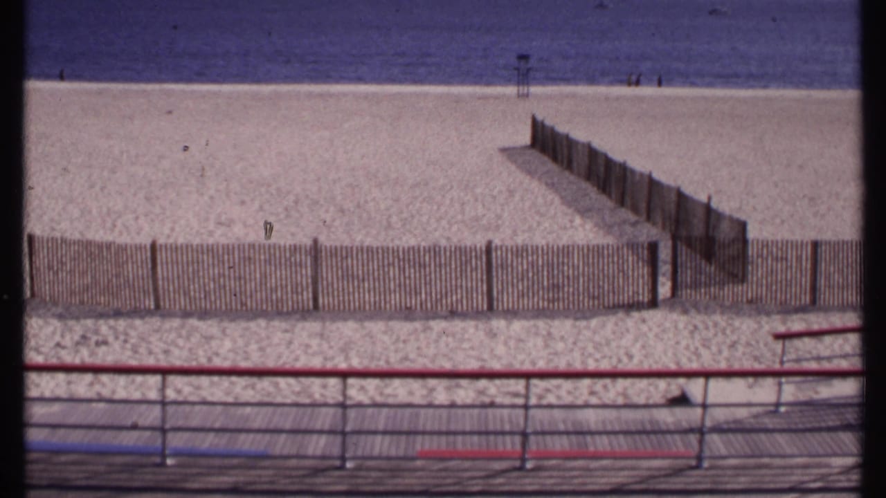 Boardwalk railing overlooks fenced sand, ocean, and a distant lifeguard stand.