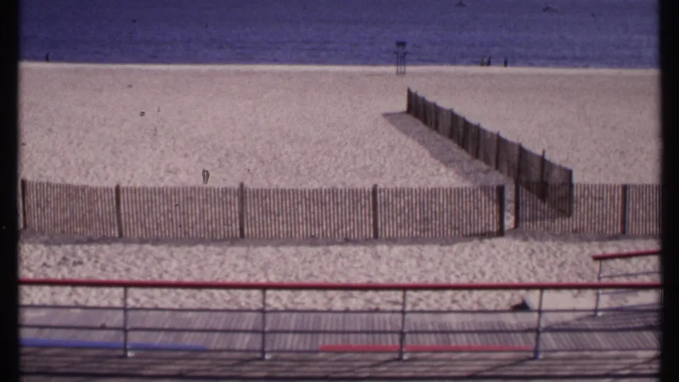 1969 New London beach view from a boardwalk, with painted railings, wooden sand fencing, ocean horizon, and a distant lifeguard stand.