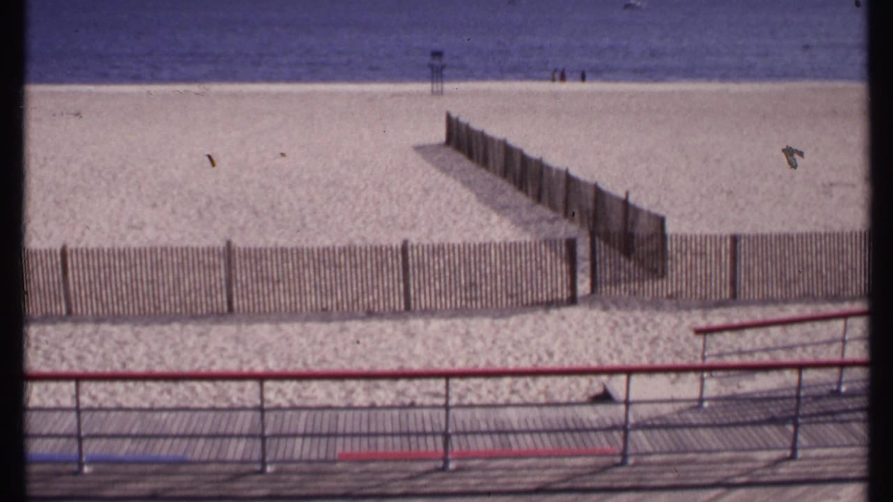 New London beach seen from the boardwalk in 1969, with sectioned wooden fencing, blue water, and small distant beachgoers near the shore.