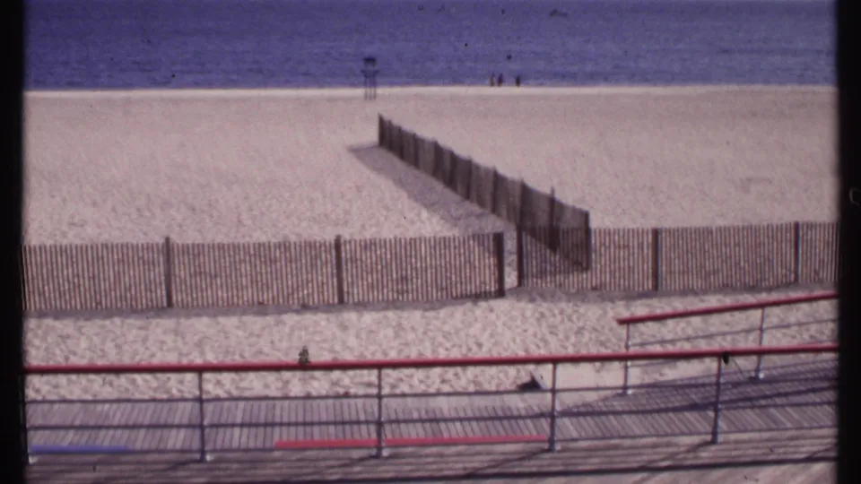Elevated 1969 view of a New London beach, showing a boardwalk rail, fenced sand sections, ocean horizon, and small figures at the waterline.
