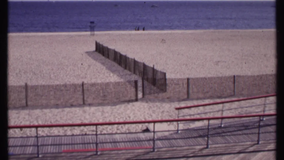1969 New London shoreline view with wooden beach divisions, red and blue boardwalk railings, bright sand, ocean, and distant people near the water.