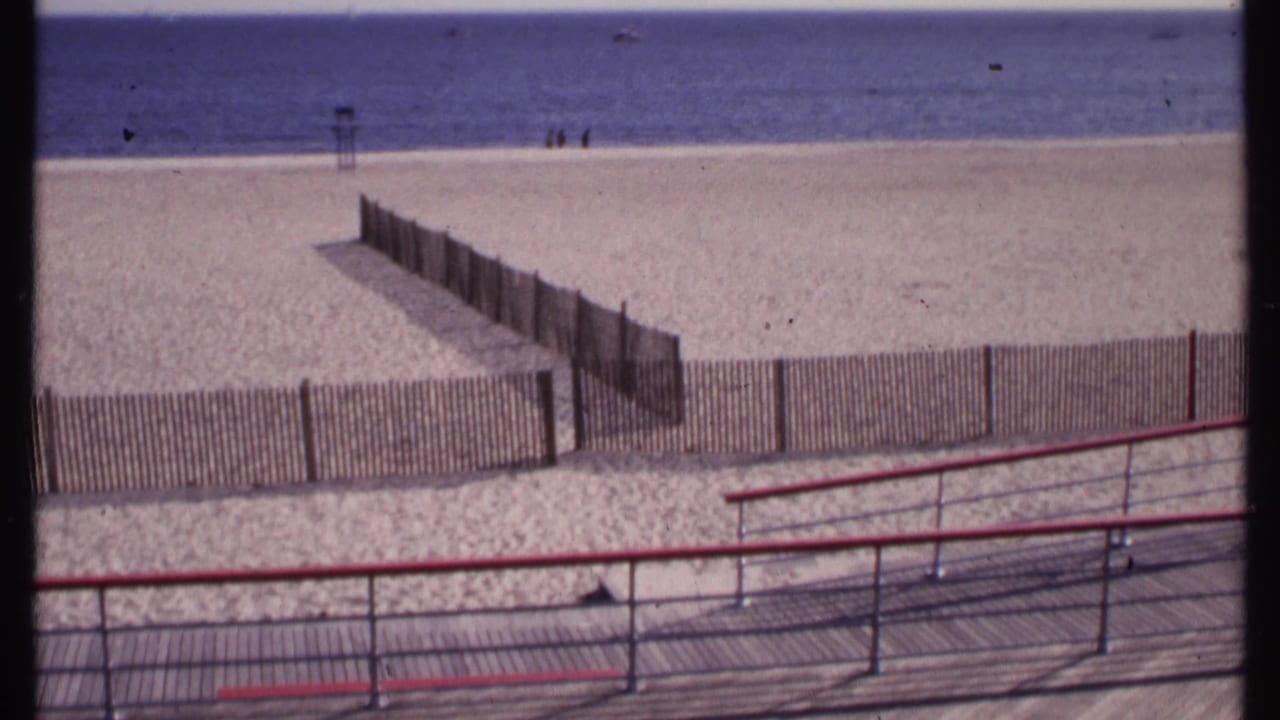 Boardwalk ramp with red rails leads down toward a fenced sandy beach and calm water.