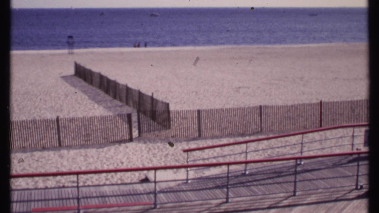 Red-railed boardwalk and slatted fencing overlook a quiet sandy beach and blue water.