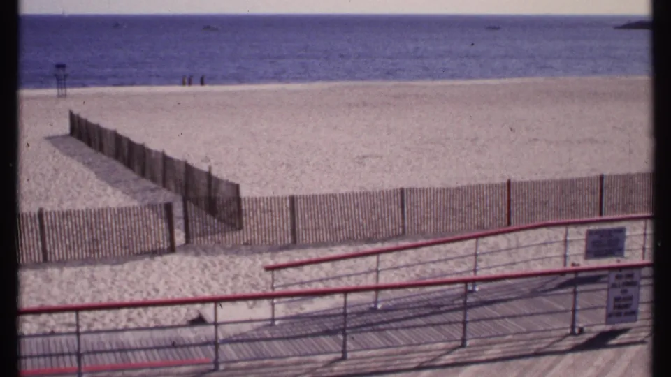 1969 home-movie frame from New London showing boardwalk railings, slatted beach fencing, broad sand, and distant beachgoers by the water.