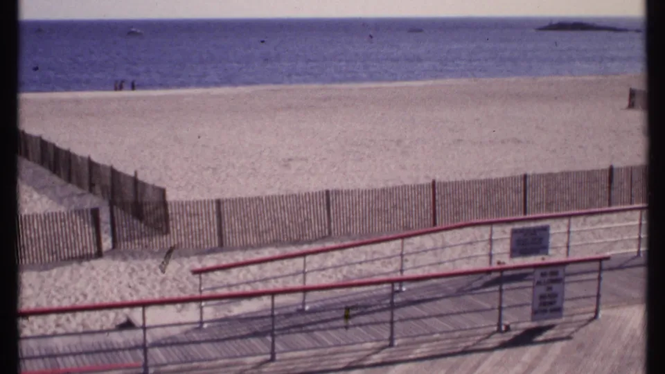 New London beach footage from 1969 with a red-railed boardwalk, fenced sand, calm blue water, distant visitors, and a small sign near the rail.