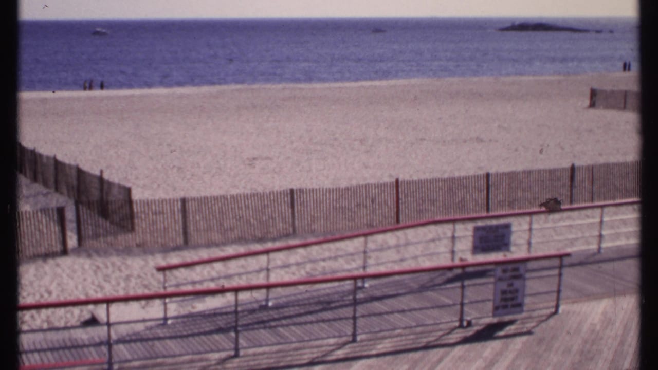 Boardwalk railing, beach fence, sand, ocean horizon, and distant figures on the shore.