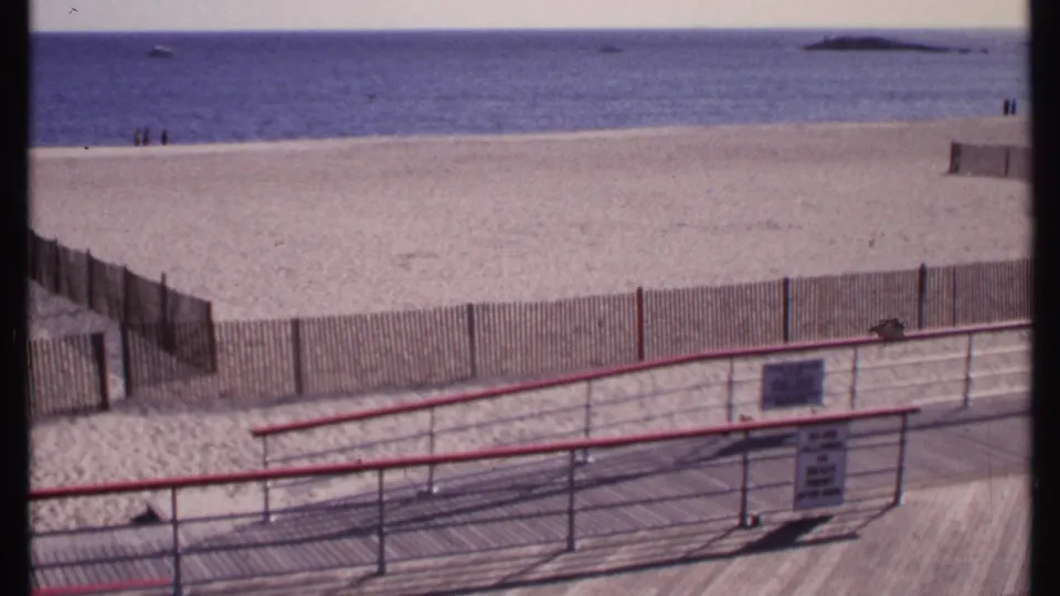 1969 New London beach view with wooden boardwalk, red-topped railing, fence, sand, and distant people near the ocean.