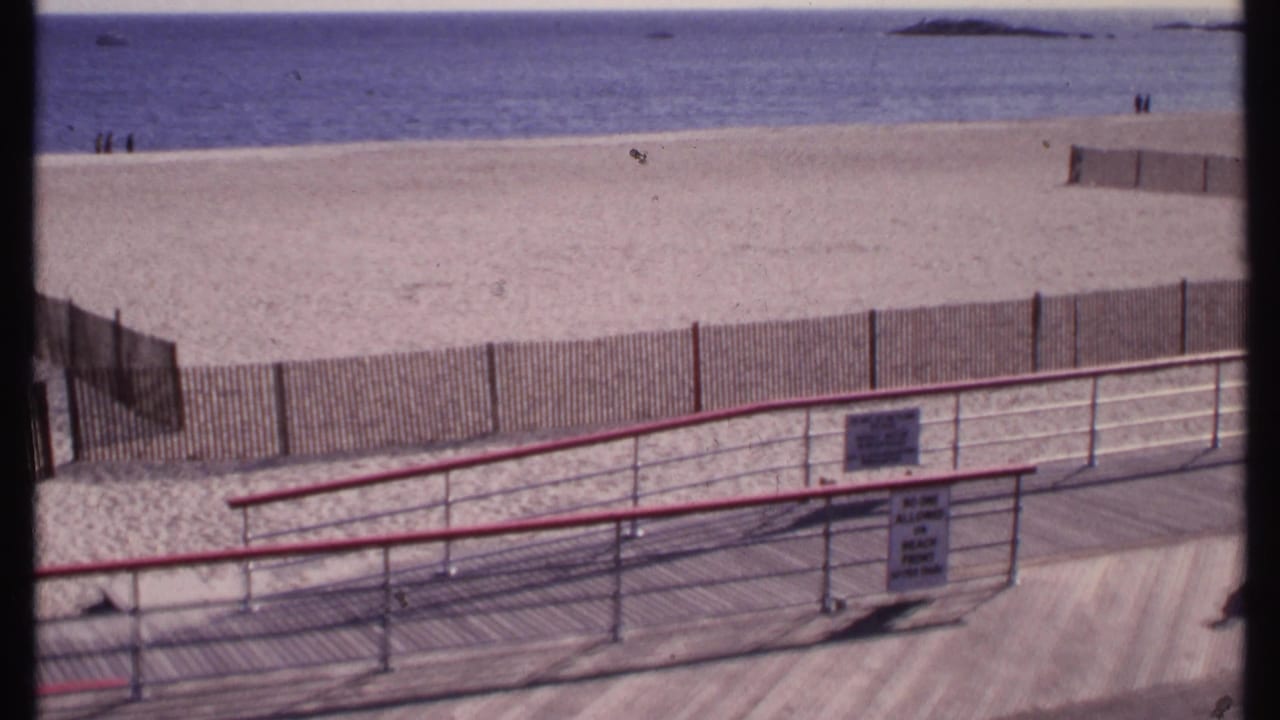 Beach, ocean, boardwalk railing, fence, and signs viewed from an elevated position.