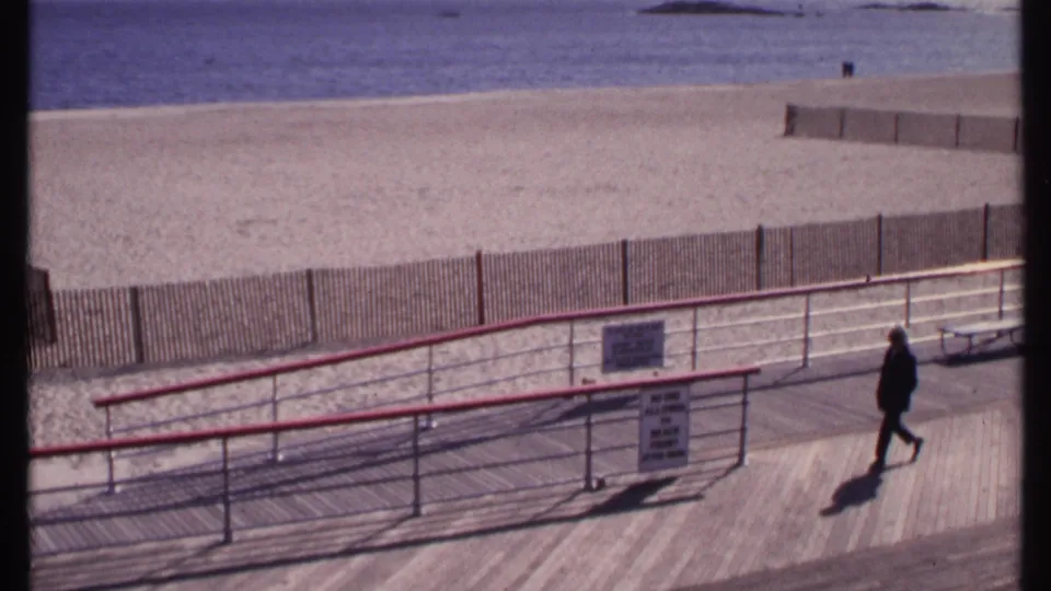 A person walks across the New London boardwalk in 1969, with beach fencing, sand, ocean, and long shadow visible.