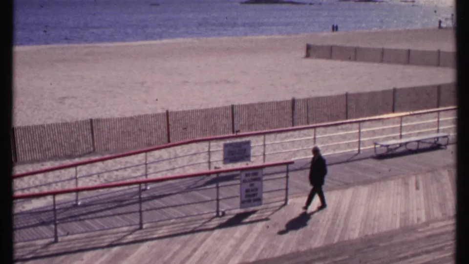 The pedestrian continues along the 1969 New London boardwalk, passing railings and signs with the beach and ocean beyond.